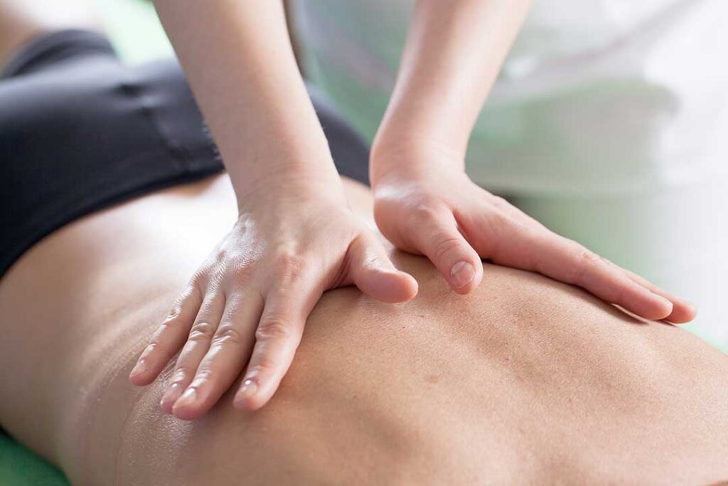 A person sitting on a bed in the morning, stretching and reaching behind their back with a look of stiffness.